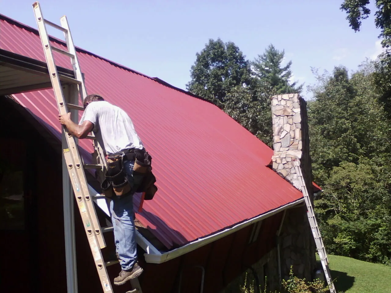 Roofer on a ladder beside a red metal roof and stone chimney — Soffit & Fascia Repair by Roofing Brothers in Lake Katrine
